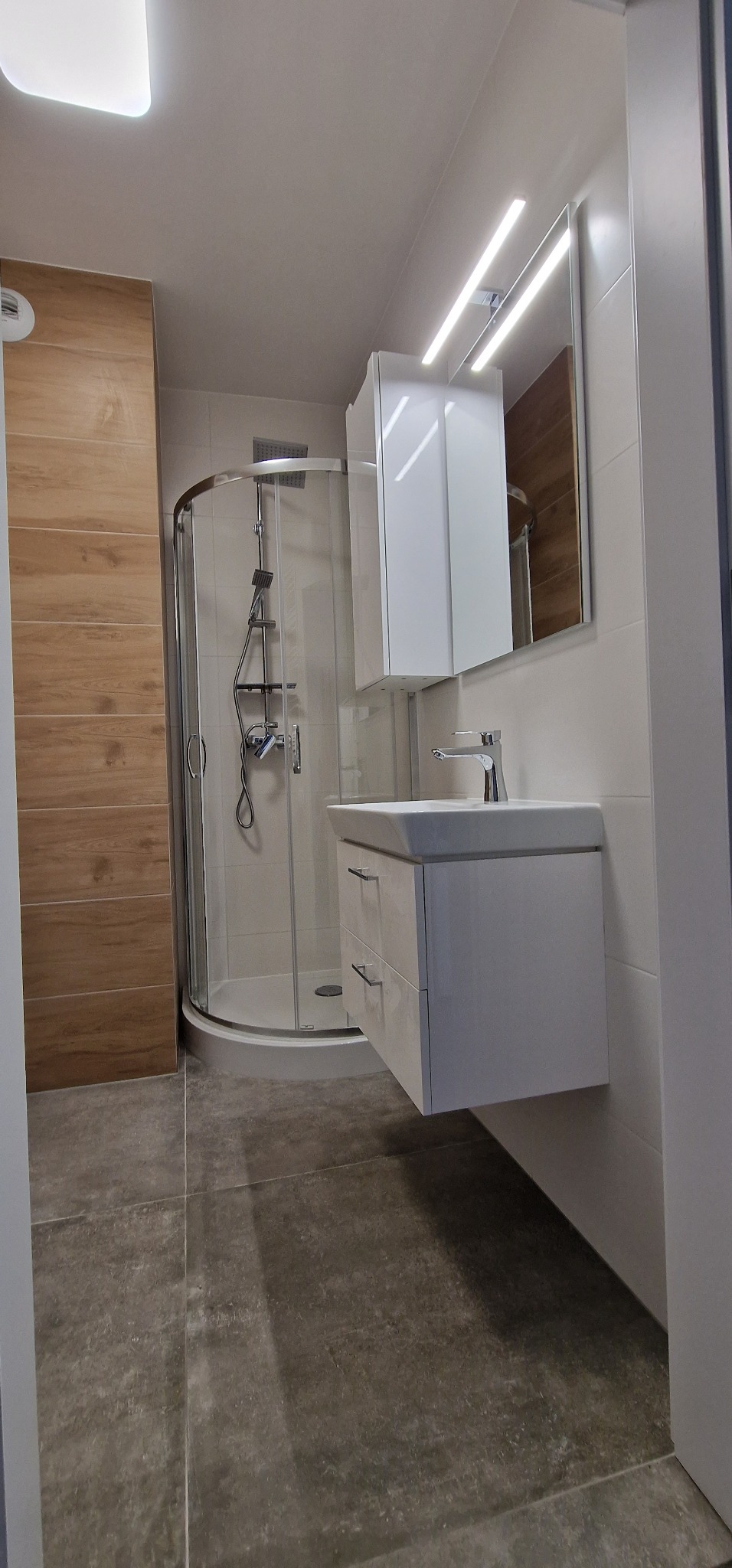 Nowoczesna łazienka z kabiną prysznicową z chromowanymi elementami, białą szafką pod umywalką z dwiema szufladami i podświetlanym lustrem, wykończona płytkami imitującymi drewno i szarą terakotą.