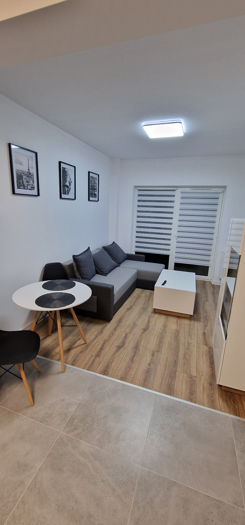 Jasne wnętrze pokoju z narożną sofą, okrągłym stołem z czarnymi podkładkami, panelami podłogowymi i szarymi płytkami podłogowymi oddzielonymi listwą, z trzema czarno-białymi obrazami na ścianie.