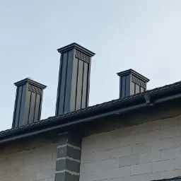 Trzy nowoczesne, grafitowe kominy wentylacyjne na dachu budynku z białego pustaka, widoczne rynny i fragment pokrycia dachowego.