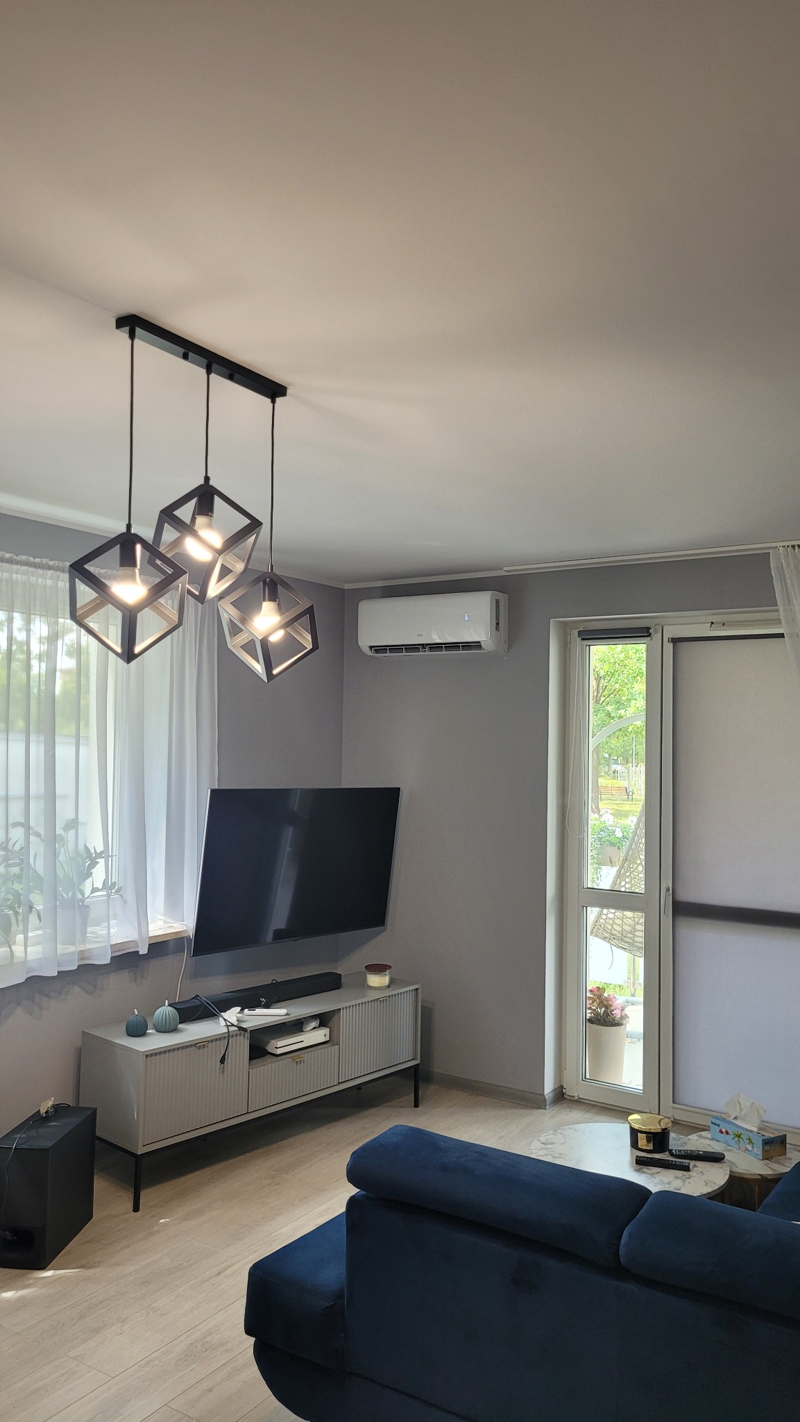 Biała jednostka klimatyzacji AUX Q-SMART zamontowana na szarej ścianie w salonie, widoczna nad telewizorem i obok okna z białymi zasłonami.