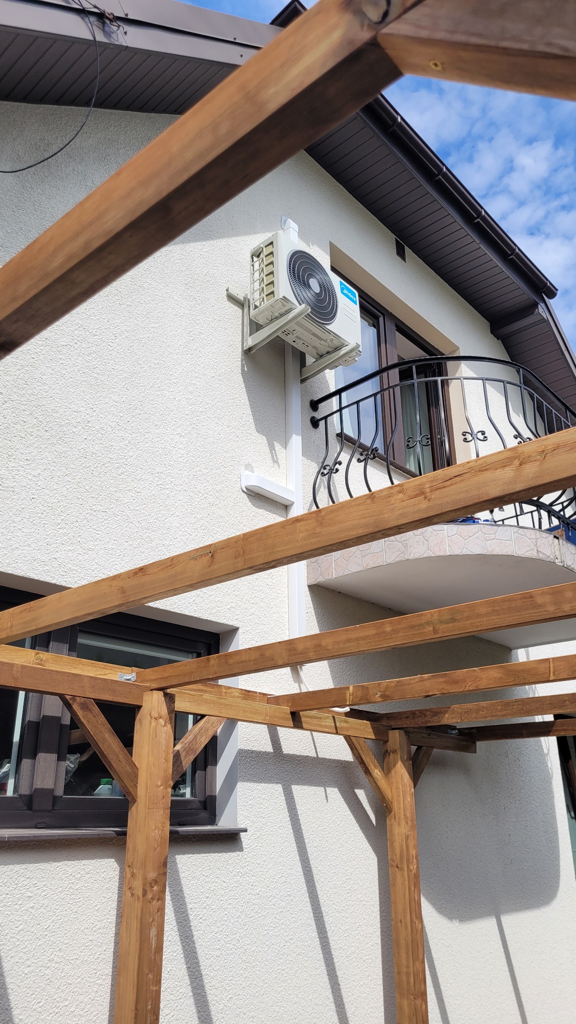 Biała jednostka zewnętrzna klimatyzacji Midea Blanc Pro 3.5kW zamontowana na elewacji domu z balkonem, widoczna spod drewnianej pergoli w budowie.