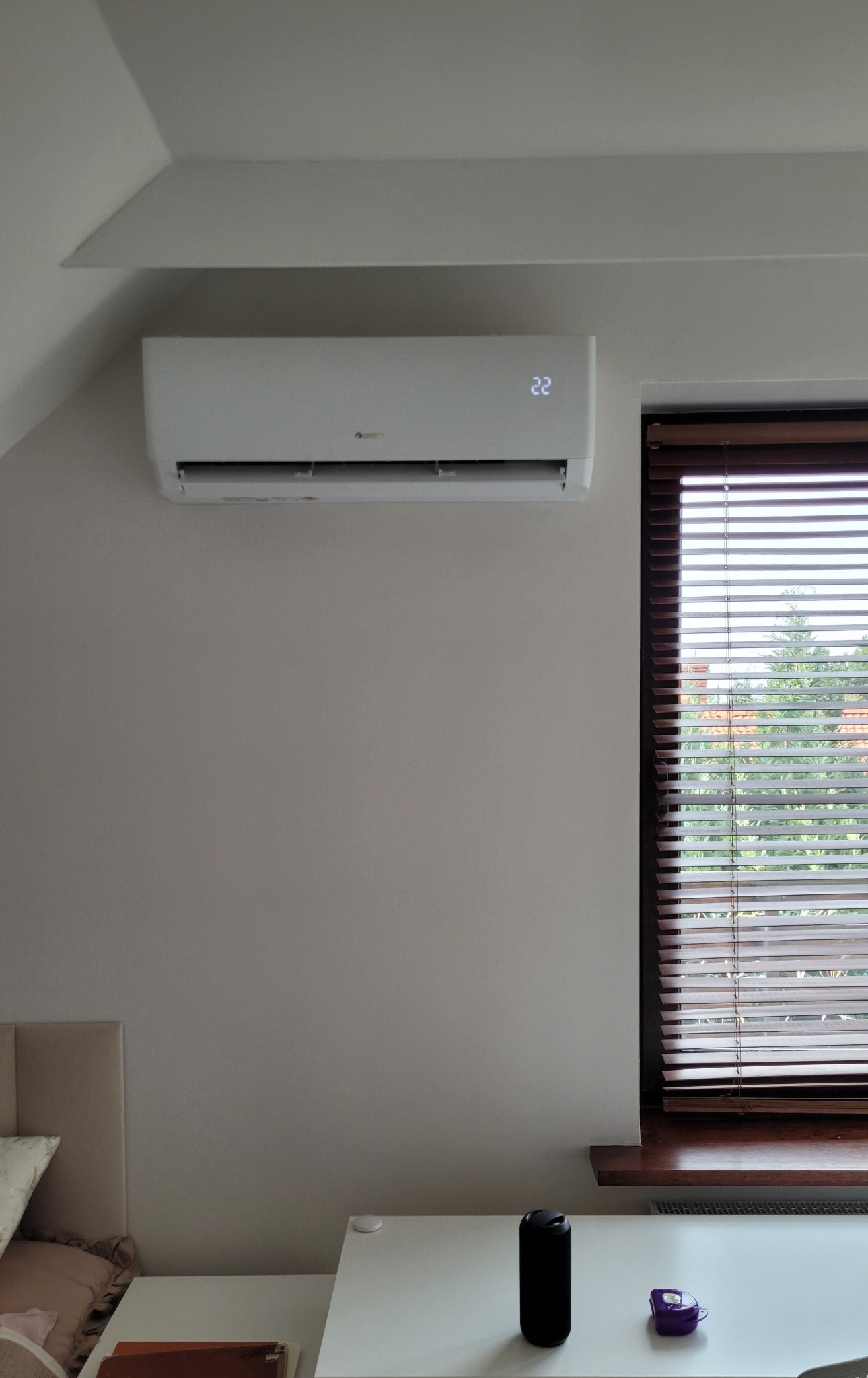Biały klimatyzator Gree Pular 3.2kW zamontowany na ścianie w jasnym pokoju z oknem w drewnianych żaluzjach, widoczny fragment łóżka i stolika.