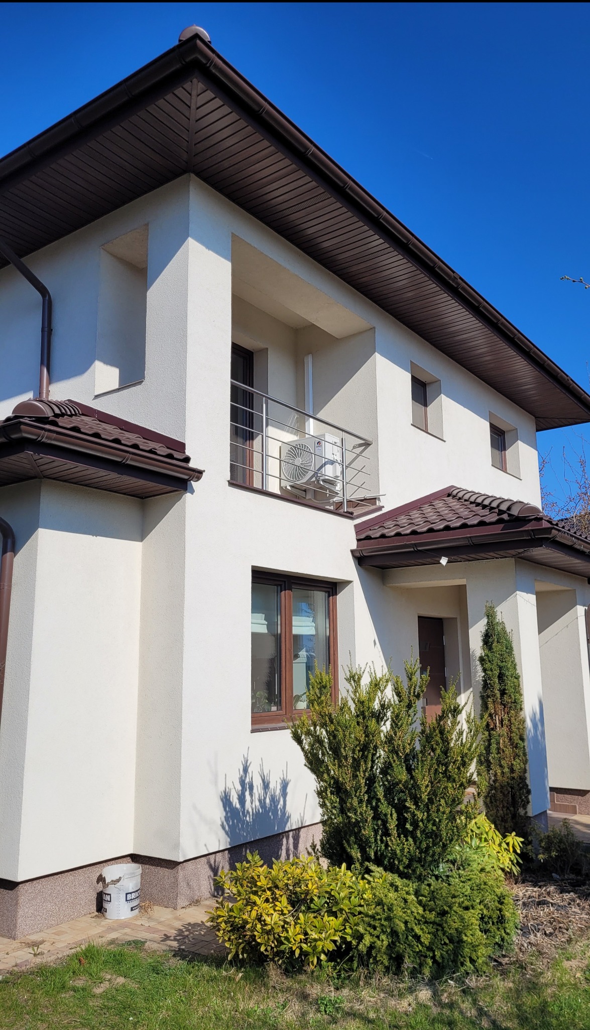 Biały dom z brązowym dachem i klimatyzatorem Gree Pular 2.5kW zamontowanym na balkonie z metalową barierką.