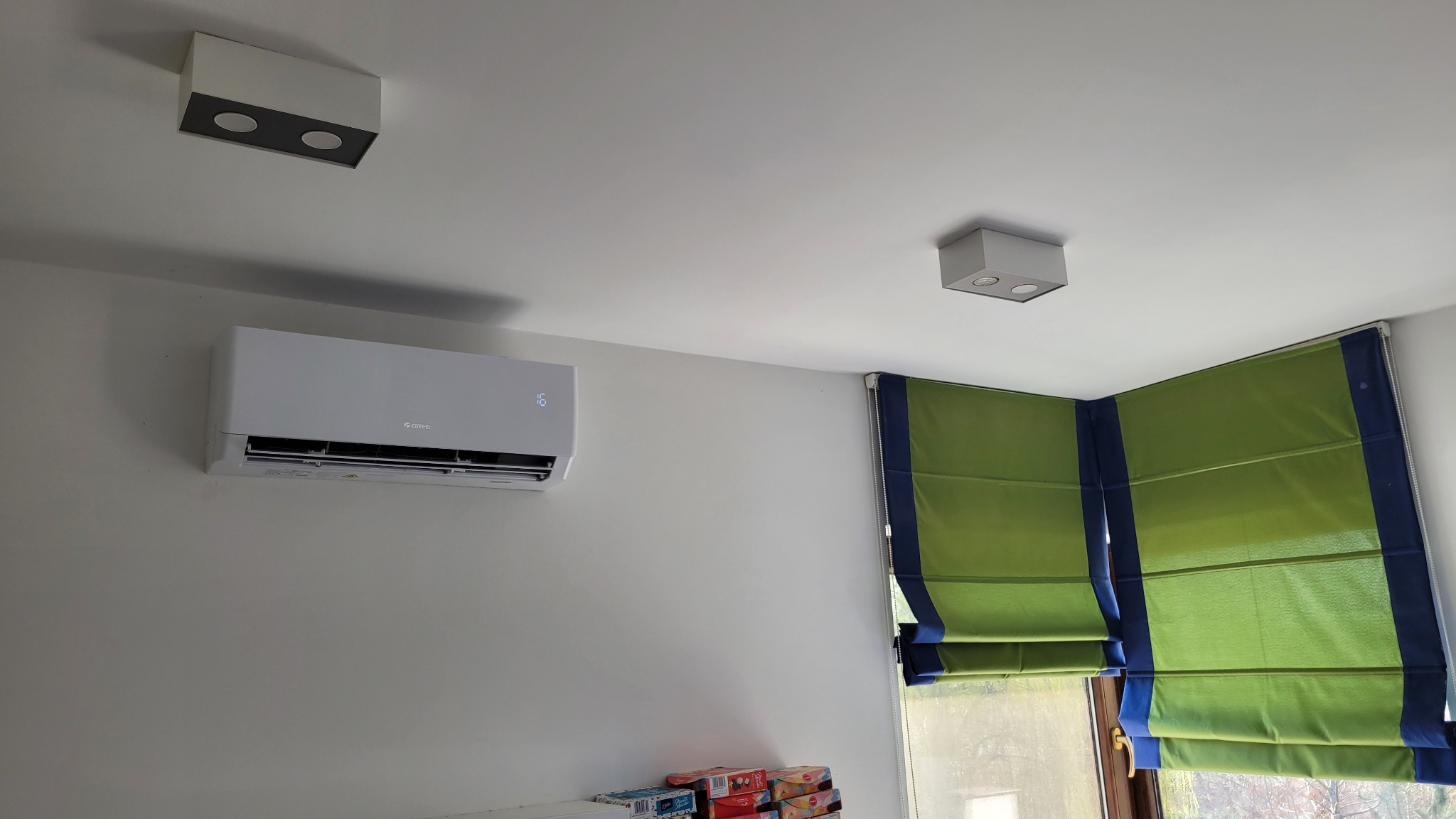 Biały klimatyzator Gree Pular 2.5kW zamontowany na ścianie w pokoju z zielonymi roletami rzymskimi i dwoma lampami sufitowymi.