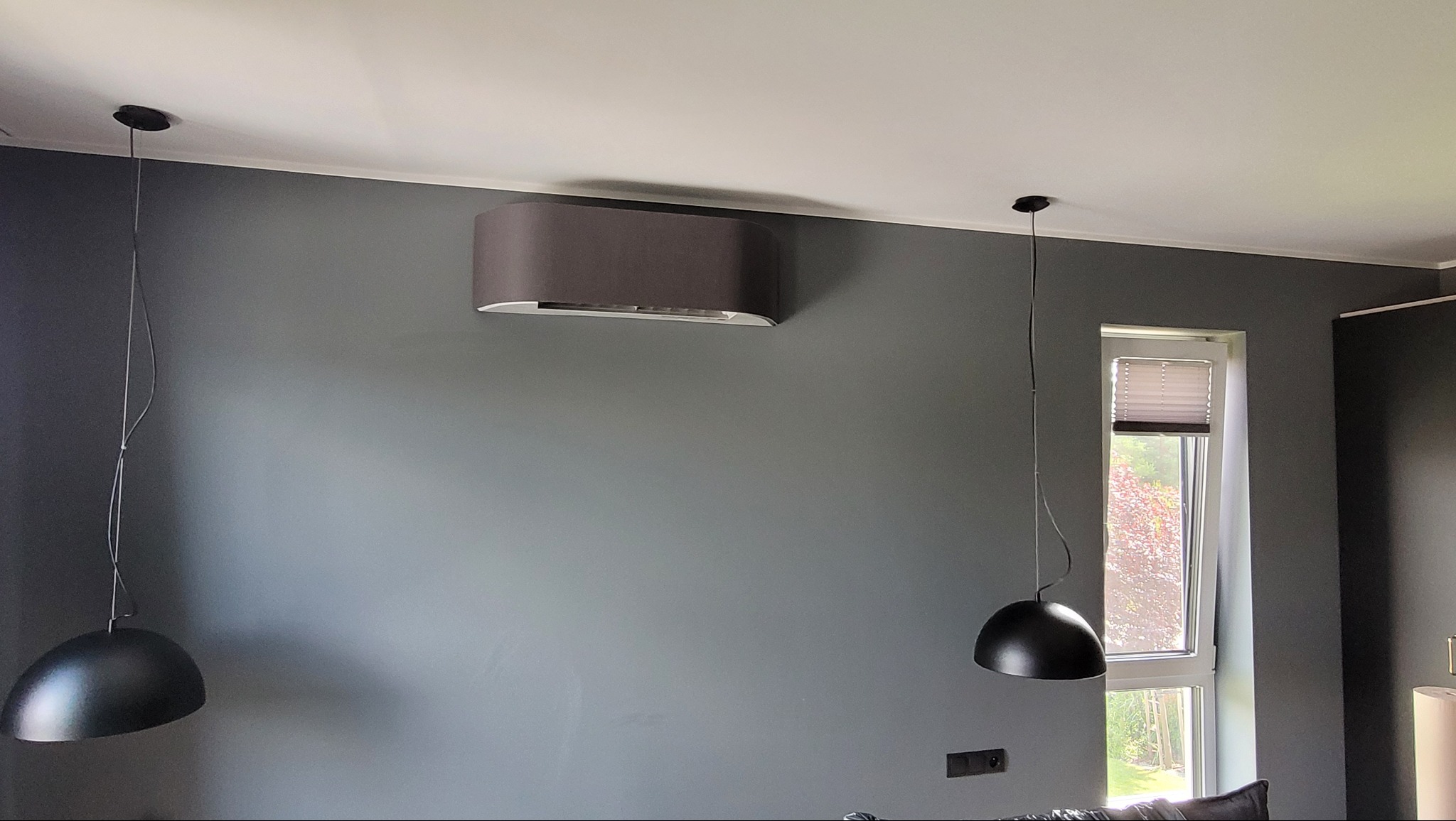 Zamontowany na szarej ścianie klimatyzator Toshiba Haori o zaokrąglonym kształcie, z widocznymi dwoma wiszącymi lampami po bokach. Nowoczesne wnętrze.