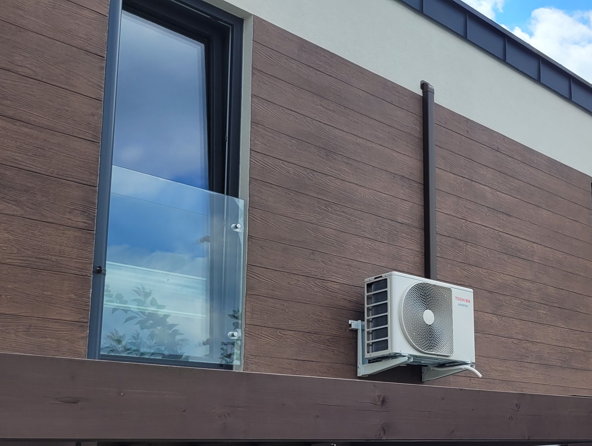 Biała jednostka zewnętrzna klimatyzatora Toshiba Haori zamontowana na brązowej elewacji budynku obok okna z balustradą. Widoczny fragment dachu i niebo.