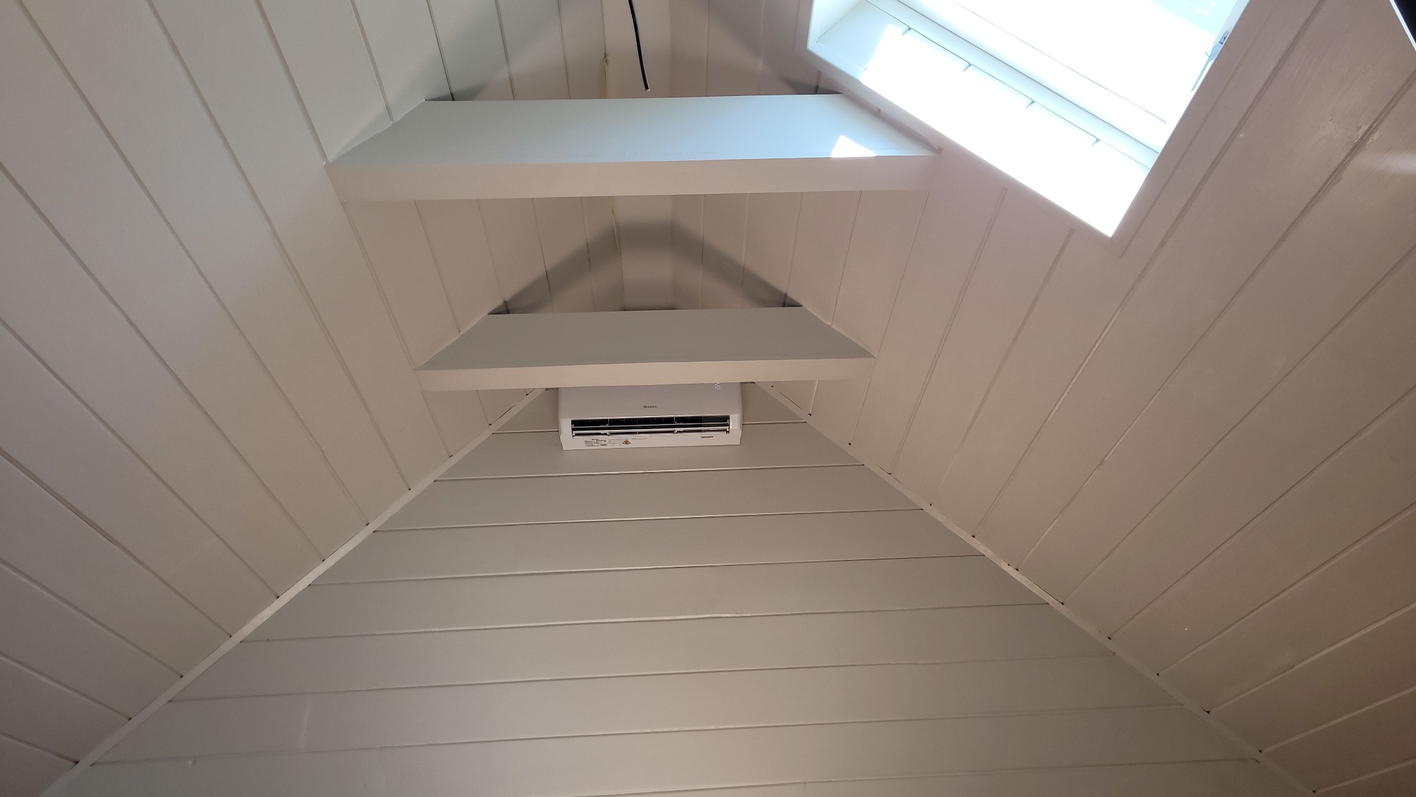 Biała klimatyzacja Gree Pular zamontowana na poddaszu z drewnianą boazerią, widok z dołu, oświetlenie naturalne z okna dachowego.