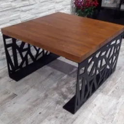 Stolik kawowy z blatem z drewna i ażurową, geometryczną podstawą z czarnego metalu, ustawiony na podłodze imitującej drewno, na tle ściany z dekoracyjnej cegły.