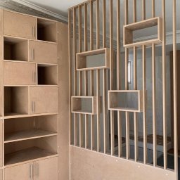 Darexpol - Surowa, niedokończona konstrukcja z płyty MDF: regał z półkami i ażurowa ścianka działowa z prostokątnymi wnękami, widoczne elementy montażowe, sufit z dekoracyjną sztukaterią.