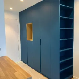 Niebieska szafa wnękowa z drewnianą wnęką i otwartymi półkami, zabezpieczona folią podłoga, fragment drewnianego blatu stołu.