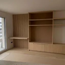 Jasne drewniane meble na wymiar w surowym stanie: szafka RTV z p&oacute;łkami i blat wkomponowany w ścianę z drewnianych listew, widok na osiedle przez duże okno.