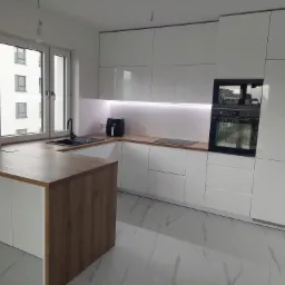 Nowoczesna, biała kuchnia z drewnianym blatem wyspowym i marmurową podłogą, oświetlona listwą LED pod szafkami wiszącymi, widok na blok mieszkalny za oknem.