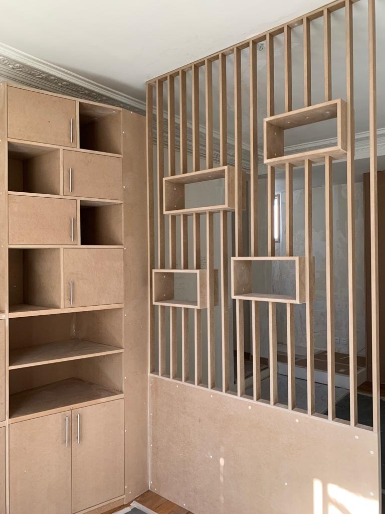 Surowa, niedokończona konstrukcja z płyty MDF: regał z półkami i ażurowa ścianka działowa z prostokątnymi wnękami, widoczne elementy montażowe, sufit z dekoracyjną sztukaterią.