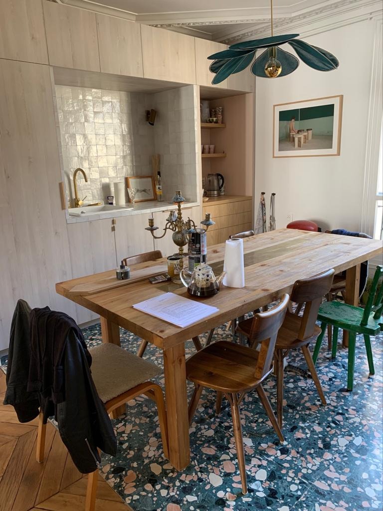 Jadalnia z drewnianym stołem, różnymi krzesłami i zabudową kuchenną w tle, widok na umywalkę z baterią w kolorze złotym i białe, kwadratowe płytki na ścianie.