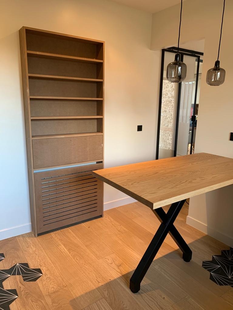 Wysoki regał z surowego drewna z częściowo osłoniętą dolną sekcją, stojący obok drewnianego stołu na metalowych nogach w kształcie litery X, na parkiecie z czarnymi, geometrycznymi wstawkami.