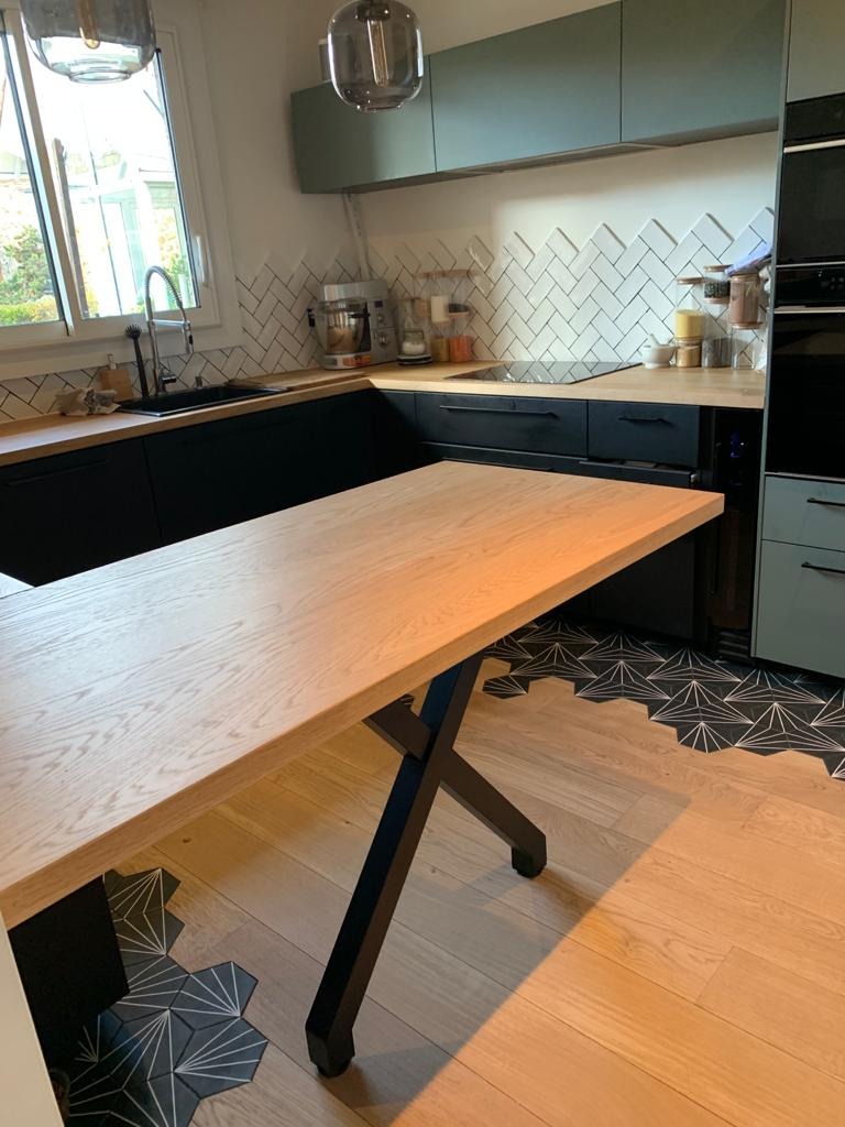 Jasna drewniana wysuwana część blatu kuchennego na czarnych metalowych nogach z kółkami, w tle czarne szafki kuchenne z drewnianym blatem i białe kafelki ułożone w jodełkę.