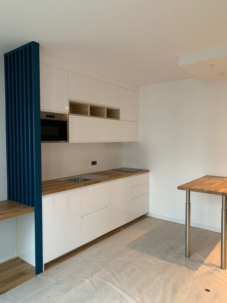 Nowoczesna kuchnia z białymi szafkami, drewnianym blatem i niebieskim ażurowym panelem, częściowo zakryta folią ochronną na podłodze.