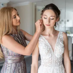 Panna młoda w białej sukni z koronkowym dekoltem, podczas zakładania kolczyków przez kobietę w błyszczącej sukience.