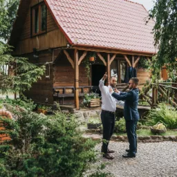 Przygotowania do ślubu przed drewnianym domem: mężczyzna w białej koszuli poprawia krawat, drugi w granatowym garniturze pomaga, w tle rustykalny dom z czerwoną dachówką i zielony ogród.