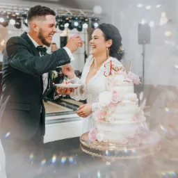 Uśmiechnięta para młoda karmiąca się tortem weselnym z dekoracjami kwiatowymi, na tle zespołu muzycznego i oświetlenia scenicznego.