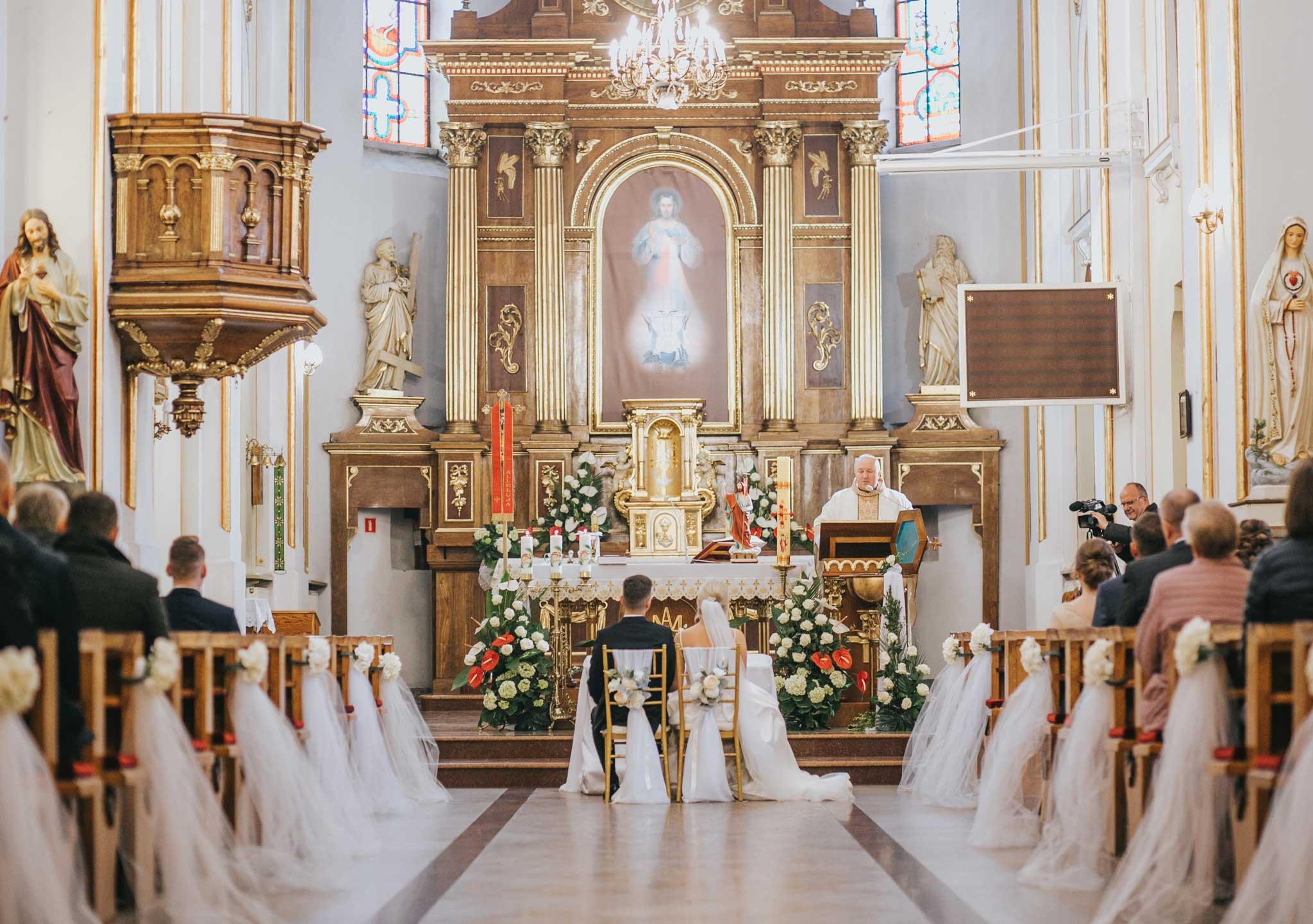 Para młoda podczas ceremonii ślubnej w bogato zdobionym kościele, ksiądz przy ołtarzu, dekoracje kwiatowe i białe tiulowe ozdoby krzeseł.