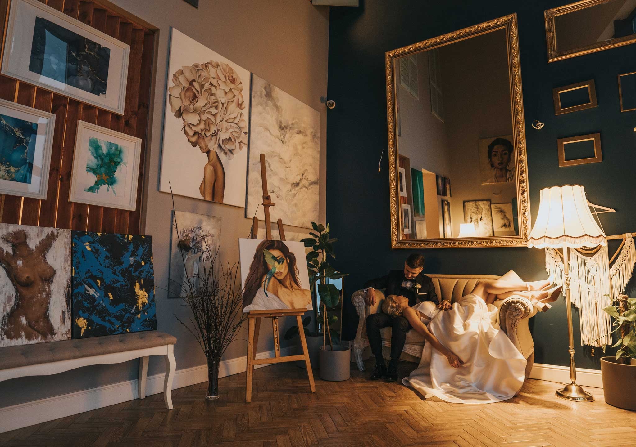 Para młoda w eleganckim wnętrzu z obrazami i lustrem w złotej ramie, panna młoda w sukni leżąca na sofie, obok sztalugi z portretem kobiety.