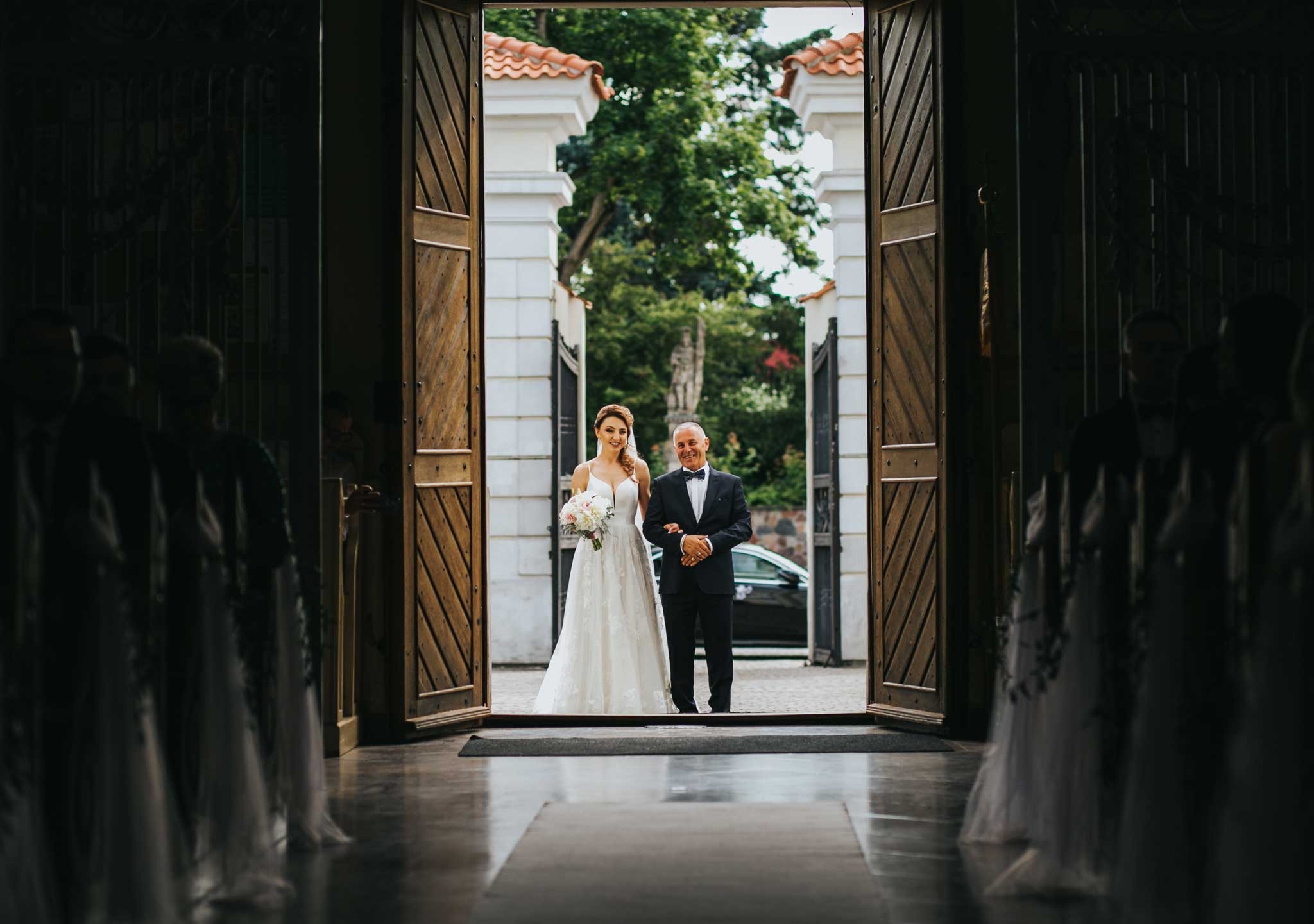 Panna młoda w białej sukni ślubnej prowadzona przez ojca w smokingu przez otwarte drewniane drzwi kościoła, widok z wnętrza kościoła, w tle zieleń i biała architektura.