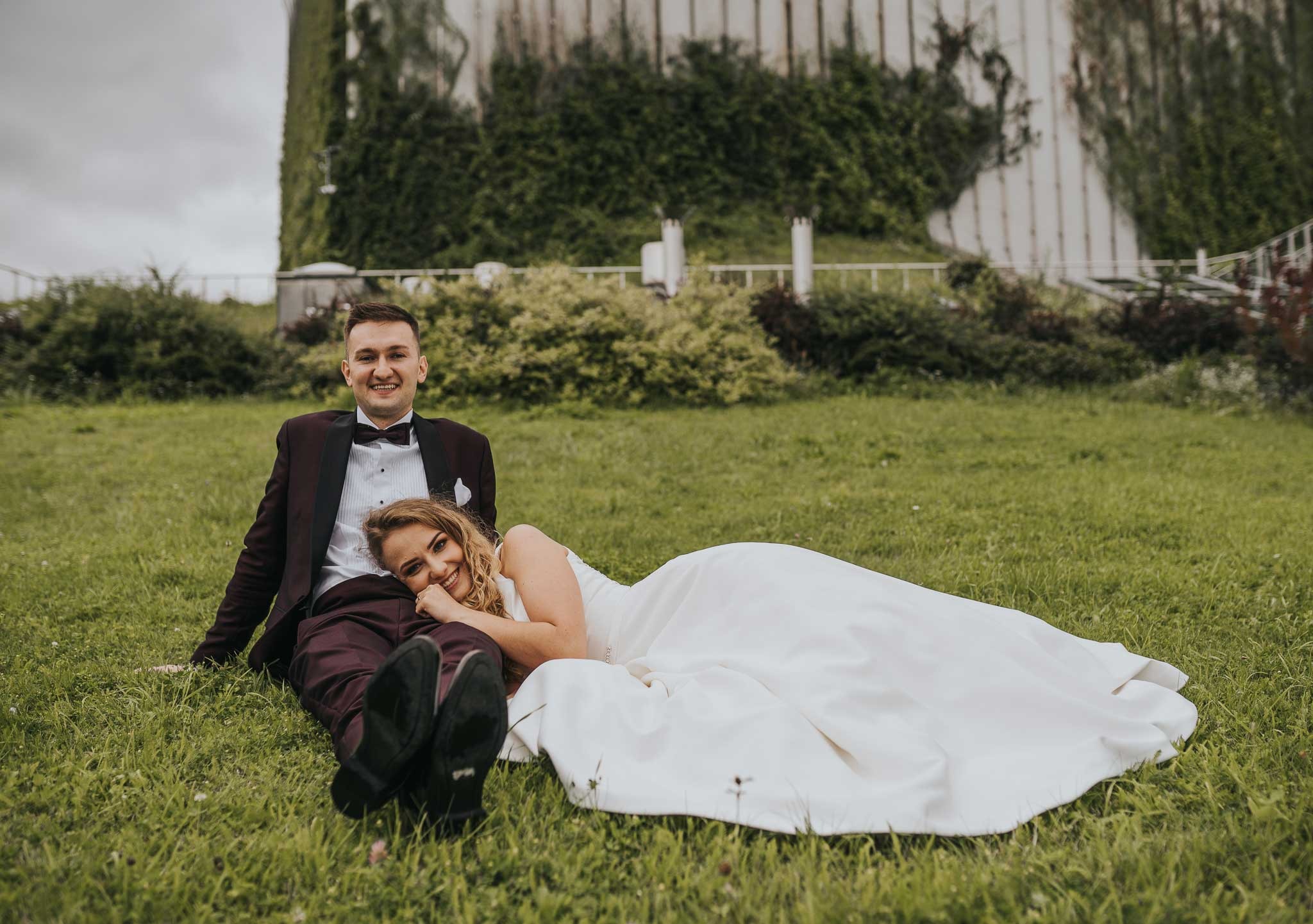 Uśmiechnięta para młoda w bordo strojach relaksuje się na trawie przed budynkiem obrośniętym bluszczem.