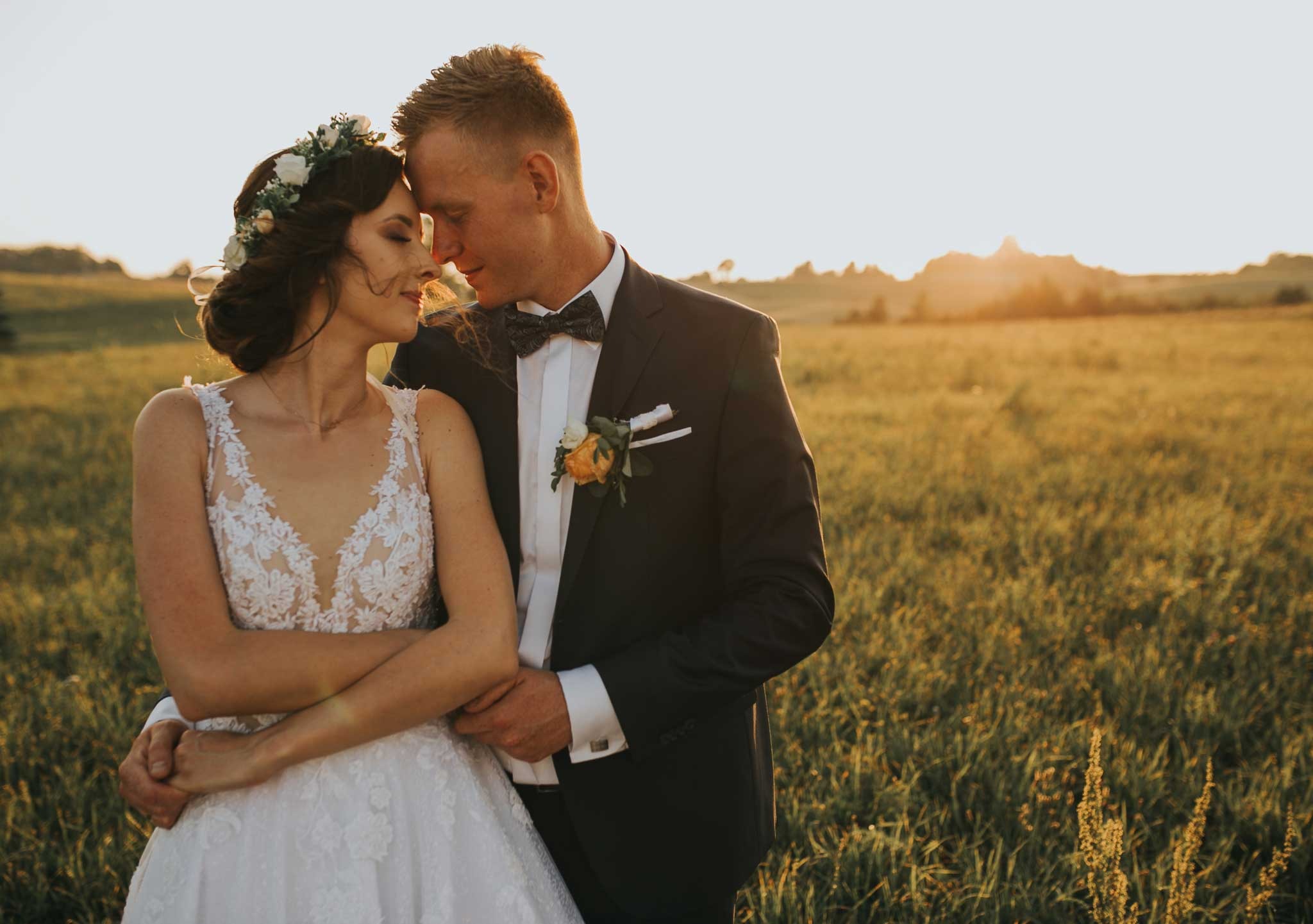 Para młoda w objęciach na tle zachodzącego słońca i pola, panna młoda w białej sukni z koronkową górą i wiankiem na głowie, pan młody w ciemnym garniturze z muszką i butonierką.