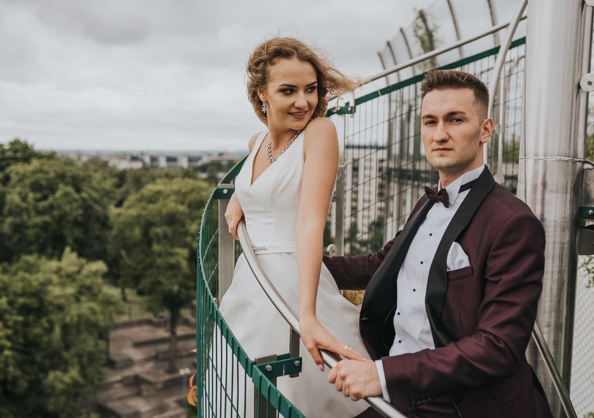 Elegancka sesja ślubna w Białymstoku: para młoda na spiralnych schodach z widokiem na miasto, panna młoda w białej sukni z biżuterią, pan młody w bordowym smokingu.