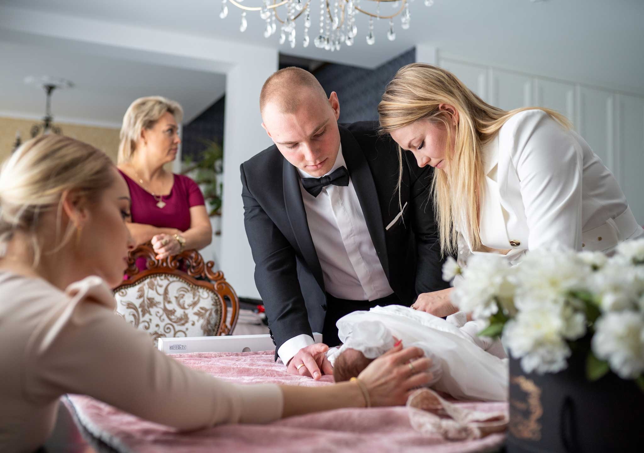 Rodzice i kobieta w beżowej bluzce delikatnie układają noworodka na różowym stole, podczas gdy inna kobieta obserwuje z tyłu w eleganckim wnętrzu.