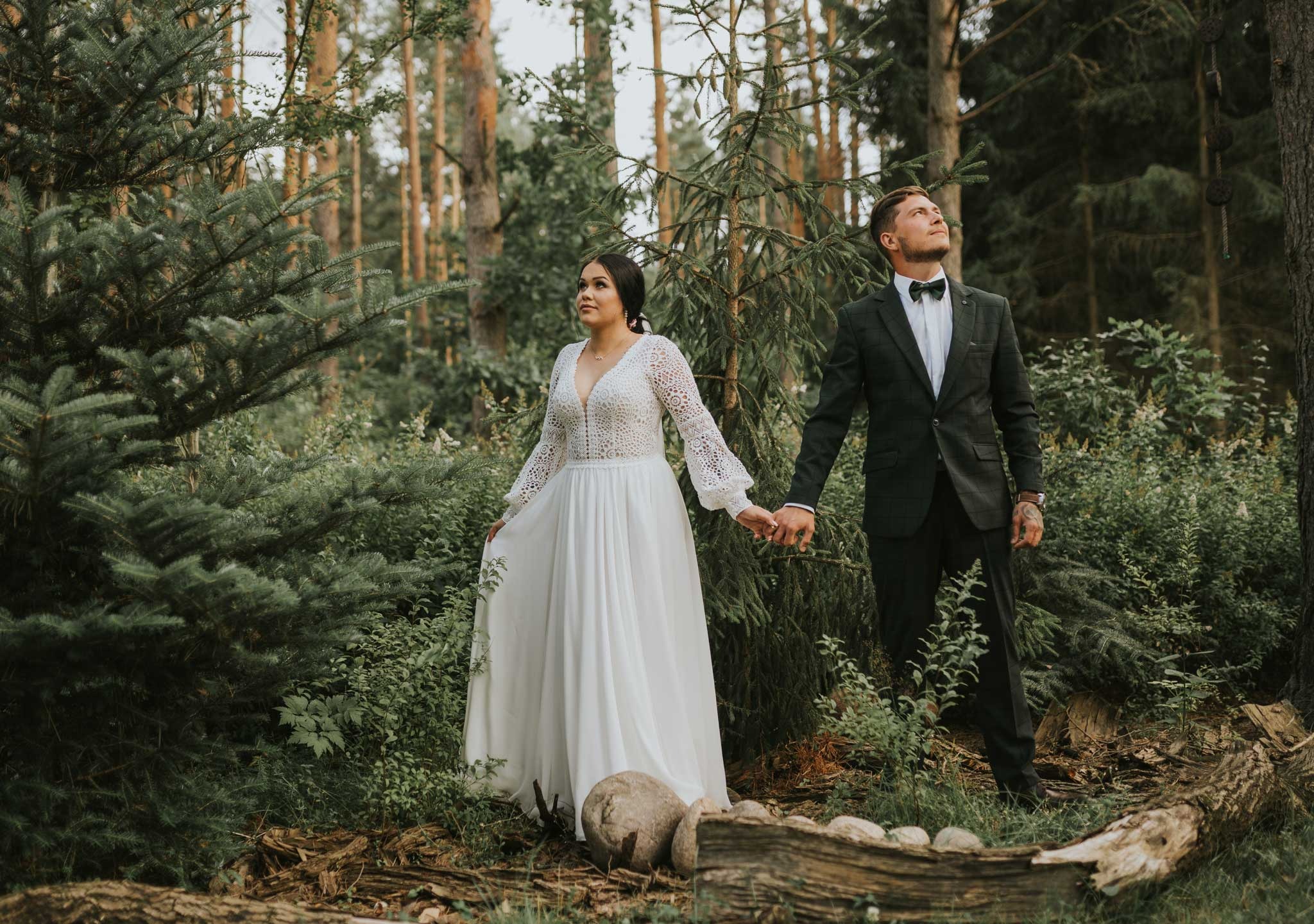 Para młoda trzymająca się za ręce w lesie, panna młoda w koronkowej sukni z długimi rękawami, pan młody w ciemnozielonym garniturze i muszce, oboje patrzą w górę, naturalne oświetlenie, leśne...