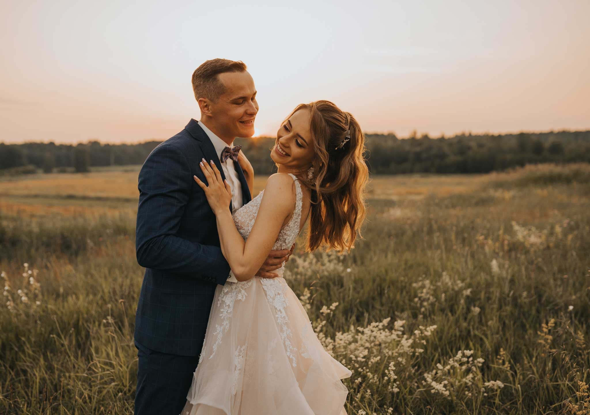 Uśmiechnięta para młoda w objęciach na łące o zachodzie słońca, panna młoda w sukni z koronkowymi aplikacjami, pan młody w granatowym garniturze w kratę i z muszką.