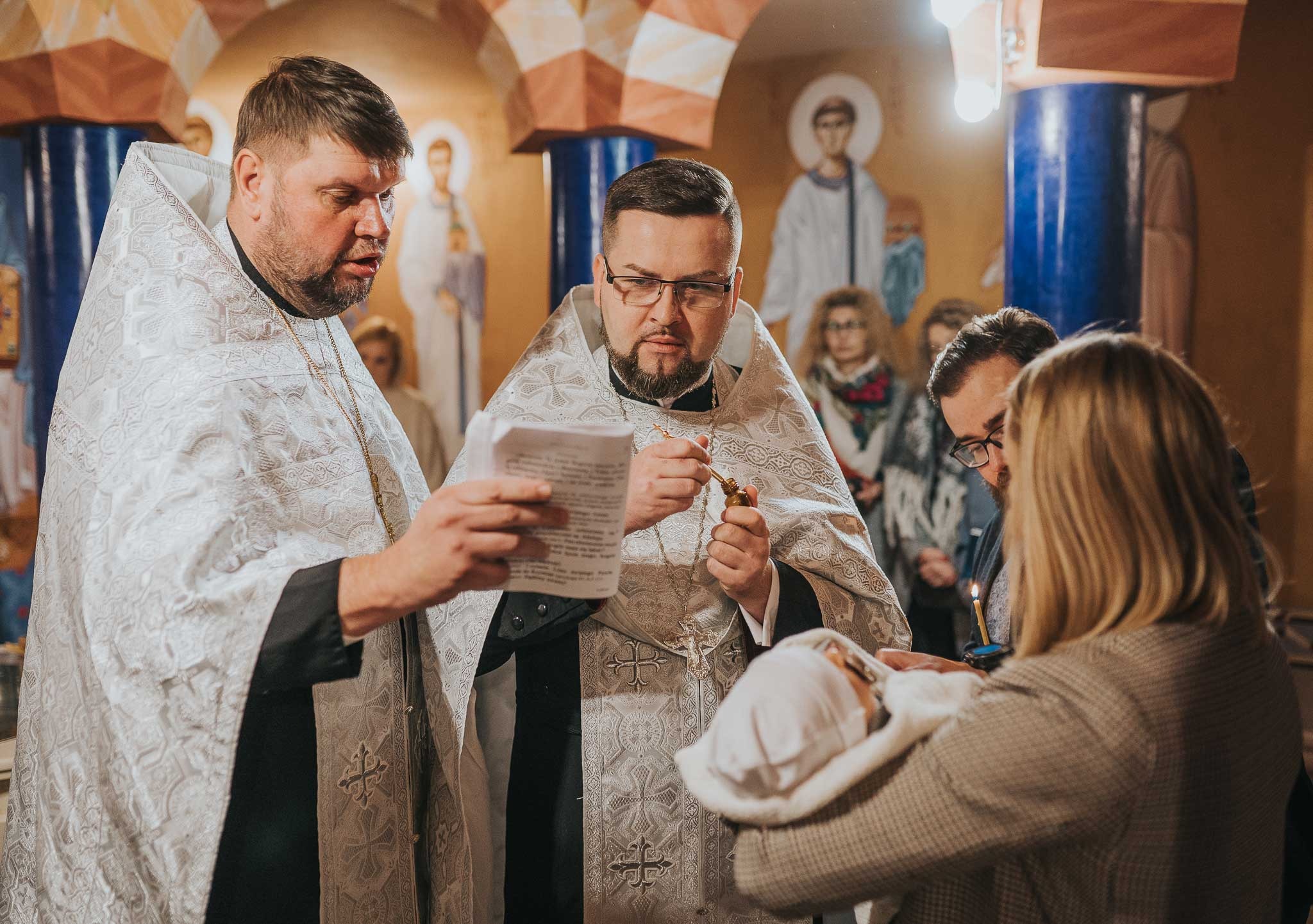 Dwóch duchownych w bogato zdobionych szatach odprawia ceremonię nad niemowlęciem trzymanym przez kobietę, w tle widać ikony i innych uczestników uroczystości.