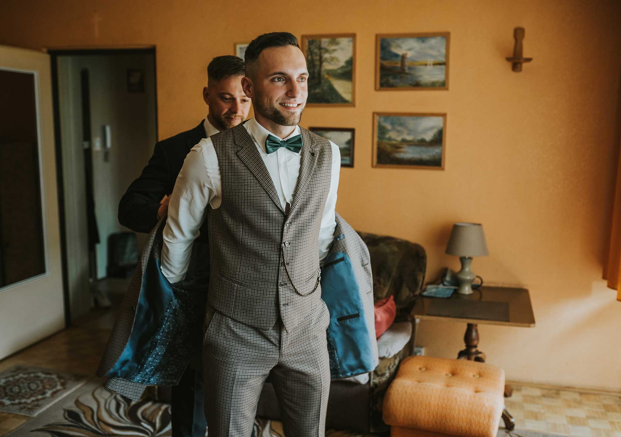 Uśmiechnięty mężczyzna w kraciastej kamizelce i zielonej muszce, pomagający mu założyć marynarkę, w tle obrazy na ścianie.