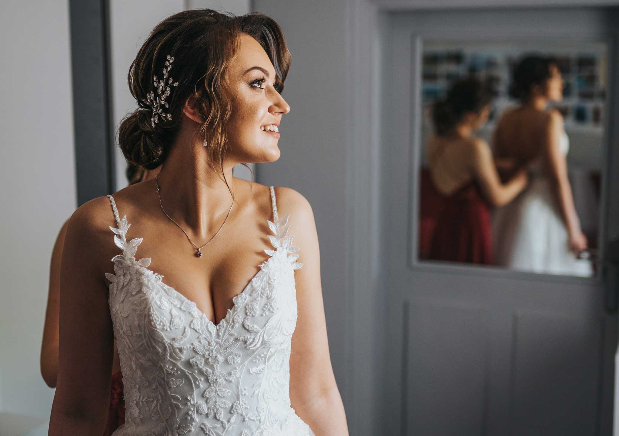 Panna młoda w białej sukni z koronkowymi zdobieniami i biżuterią we włosach, uśmiechnięta, przygotowywana do ślubu przez druhnę widoczną w odbiciu lustrzanym.