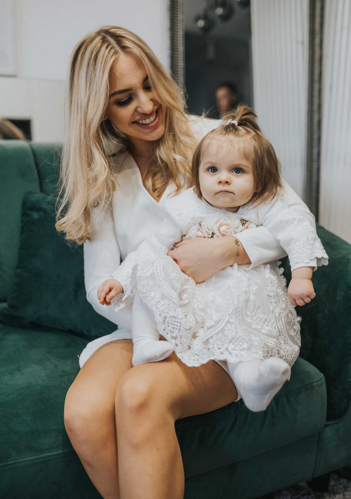 Uśmiechnięta blondynka trzyma na kolanach niemowlę w białej koronkowej sukience z różowymi aplikacjami na tle zielonej welurowej kanapy.