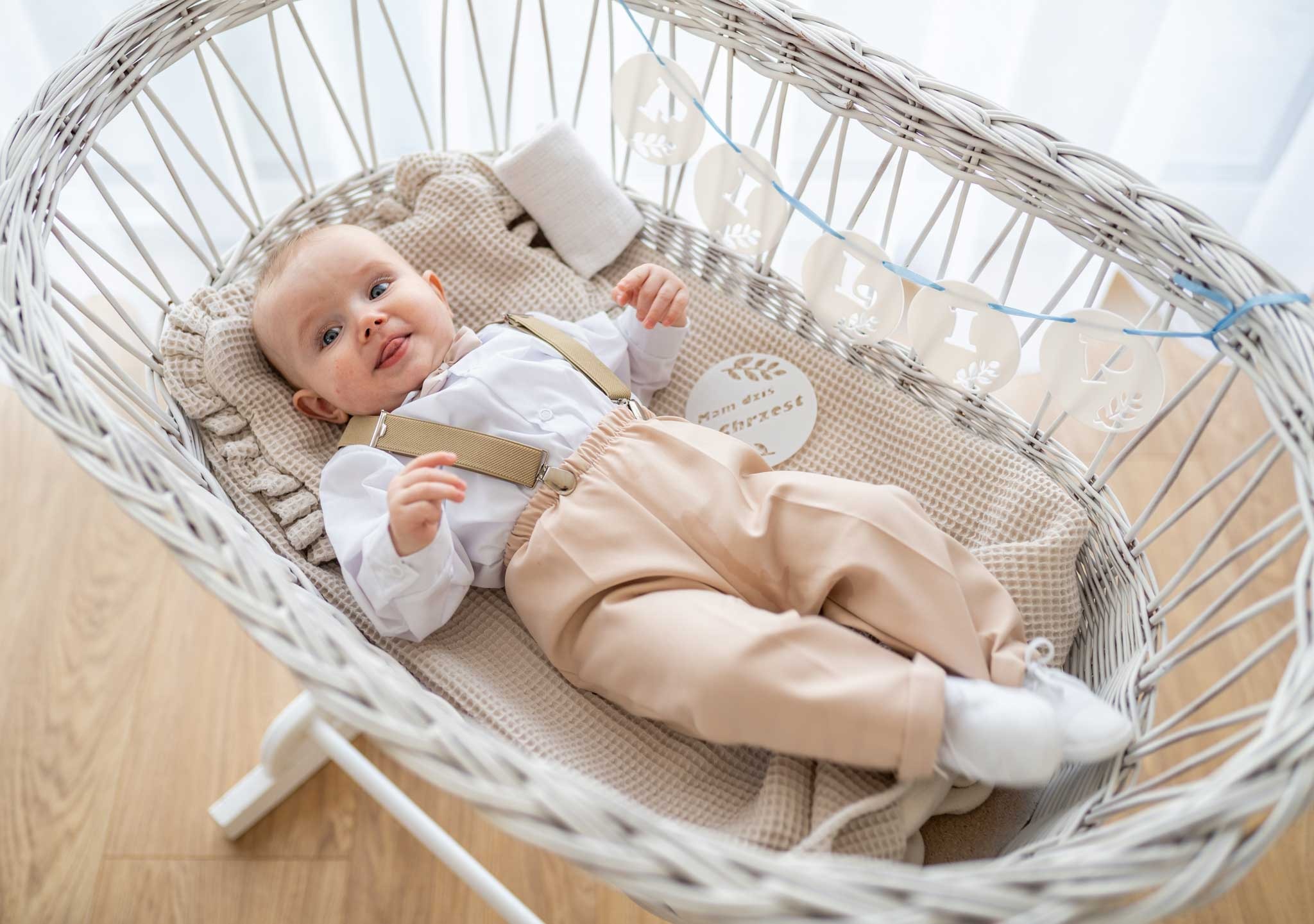 Uśmiechnięty niemowlak w białej koszuli i beżowych spodniach z szelkami leży w wiklinowym koszu, ozdobionym girlandą z napisem 'Mam dziś Chrzest'.