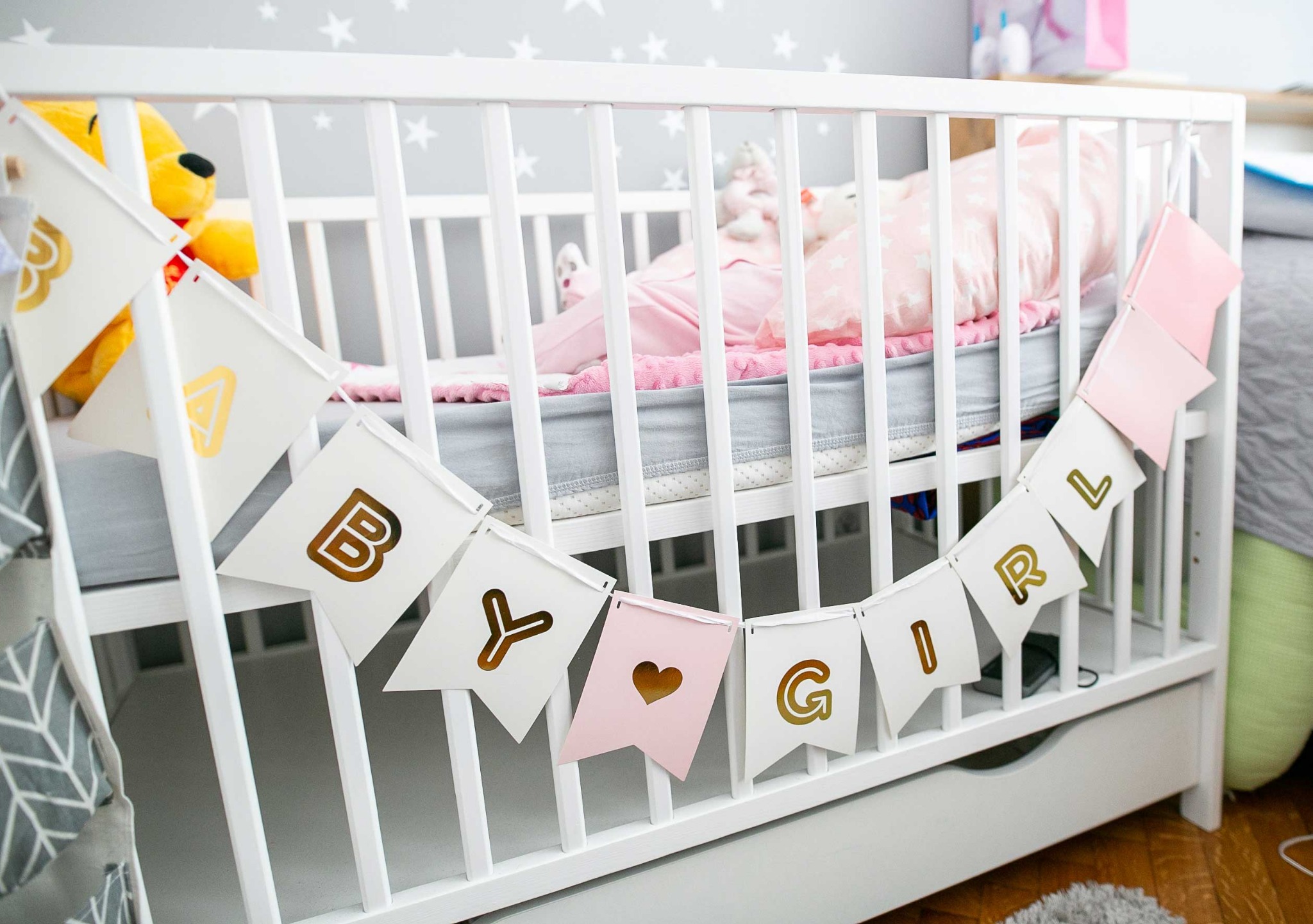 Białe łóżeczko dziecięce z dekoracyjnym banerem 'Baby Girl' w odcieniach bieli, różu i złota, pluszowy miś wystający zza barierki, pokój z szarą tapetą w gwiazdki w tle.