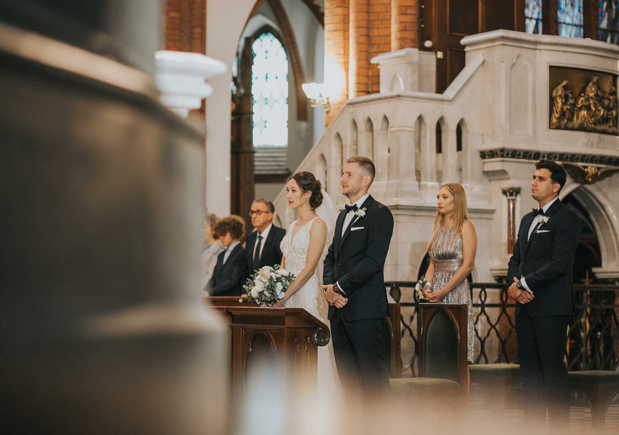 Para młoda podczas ceremonii ślubnej w kościele z ceglanymi ścianami i witrażami w tle, w otoczeniu gości ubranych w eleganckie stroje.