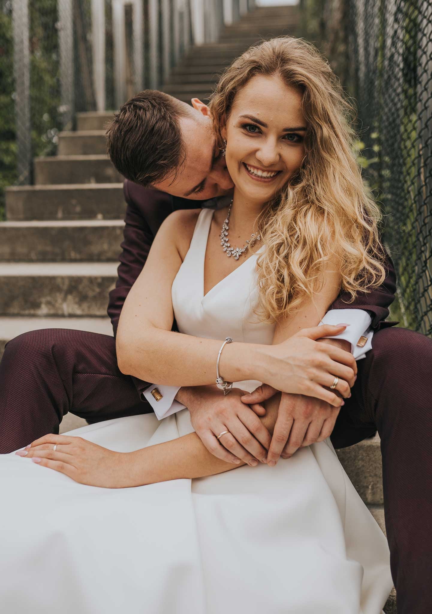 Uśmiechnięta panna młoda w białej sukni i z blond lokami, obejmowana przez pana młodego w bordowym garniturze na betonowych schodach z metalową siatką w tle.