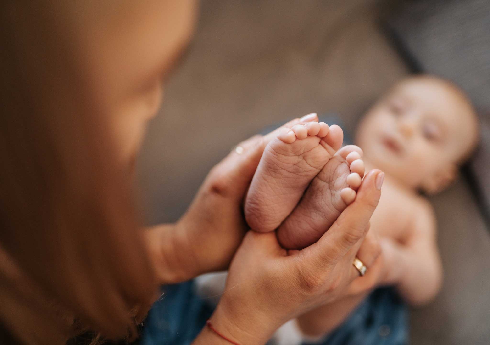 Drobne stópki noworodka w dłoniach matki, delikatne światło, pierścionek na palcu kobiety, rozmyte tło z leżącym dzieckiem.