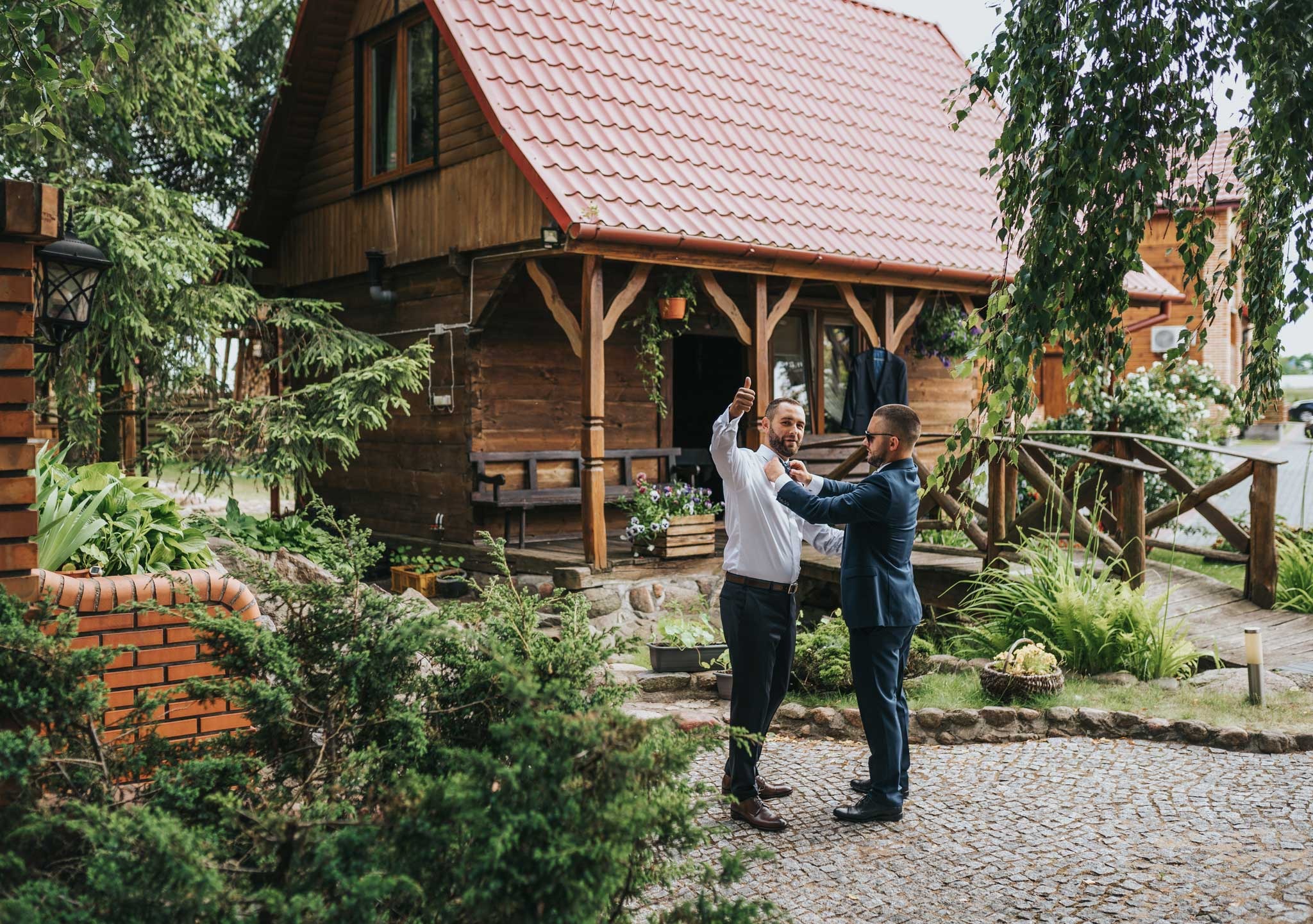 Przygotowania do ślubu przed drewnianym domem: mężczyzna w białej koszuli poprawia krawat, drugi w granatowym garniturze pomaga, w tle rustykalny dom z czerwoną dachówką i zielony ogród.