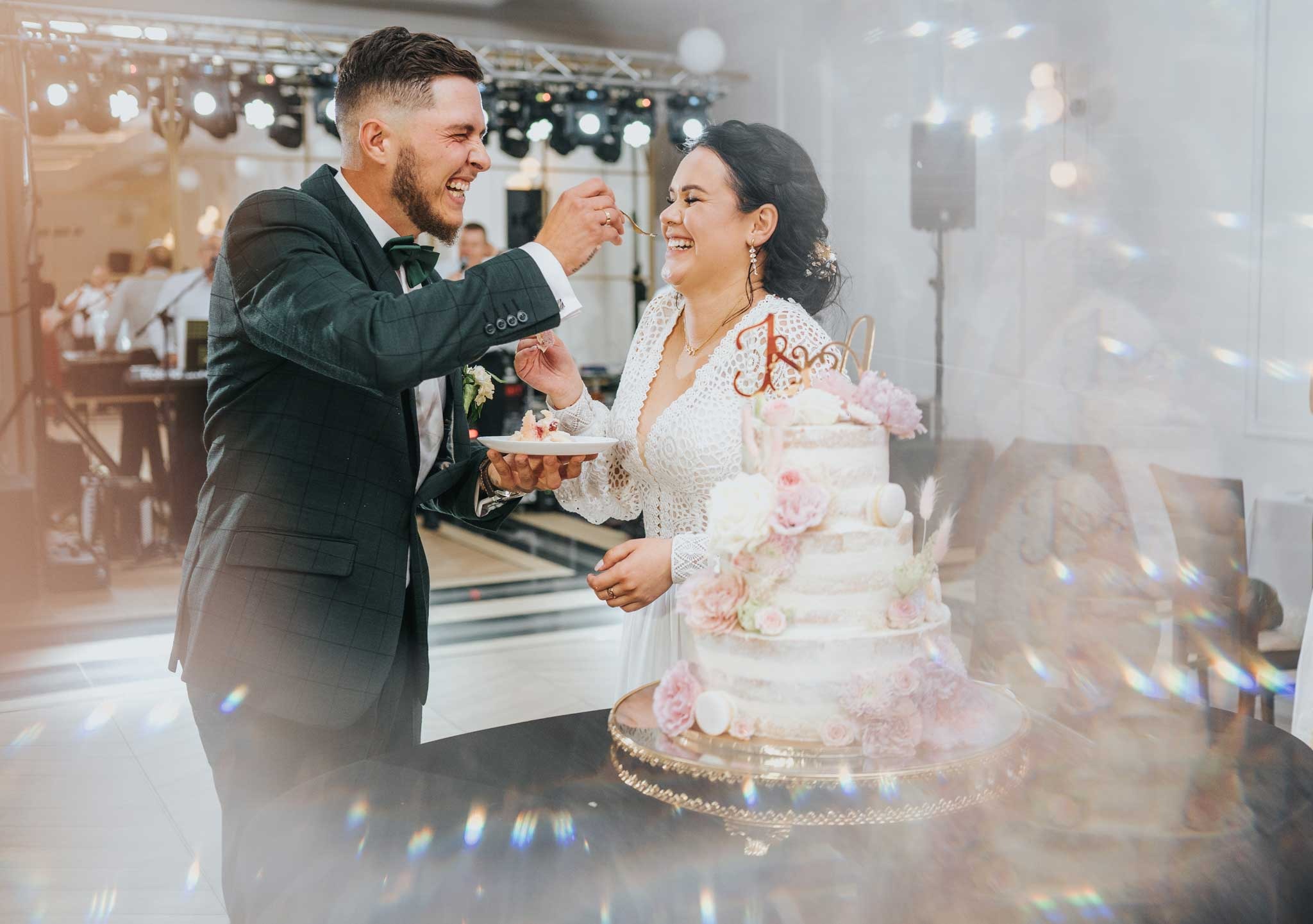 Uśmiechnięta para młoda karmiąca się tortem weselnym z dekoracjami kwiatowymi, na tle zespołu muzycznego i oświetlenia scenicznego.