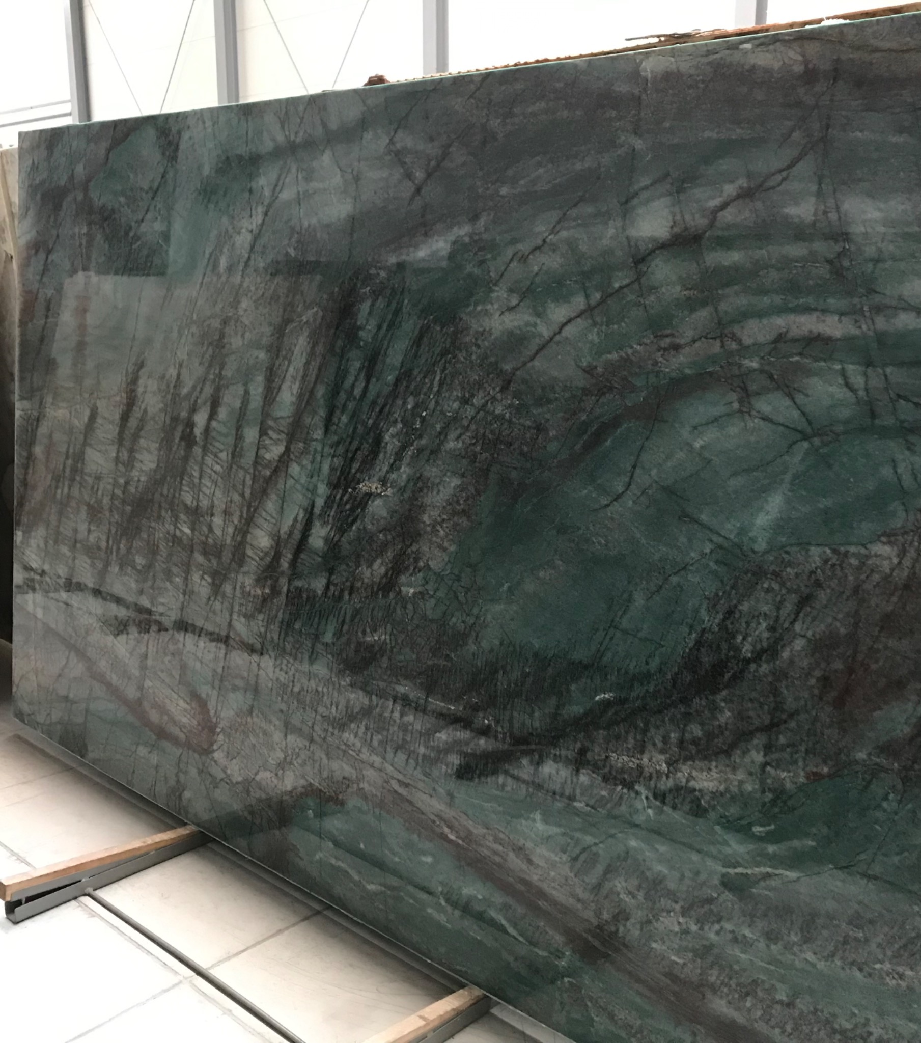 Duża płyta zielonego marmuru z wyraźnym, ciemnym żyłkowaniem, oparta na metalowej konstrukcji w magazynie.