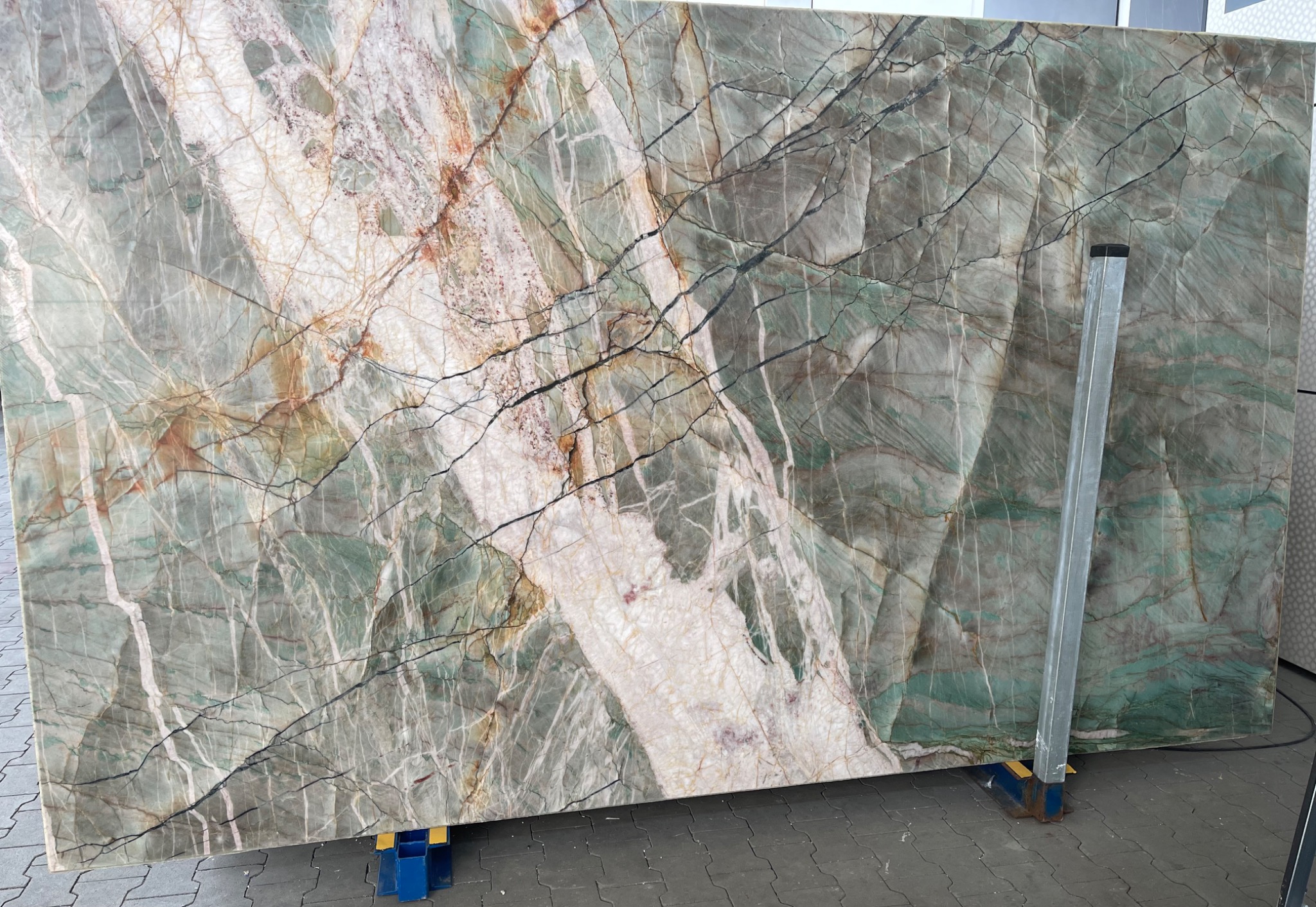 Duża płyta zielonego kamienia z białymi i brązowymi żyłami, zabezpieczona metalowym wspornikiem, prezentowana przez firmę z Mińska Mazowieckiego.