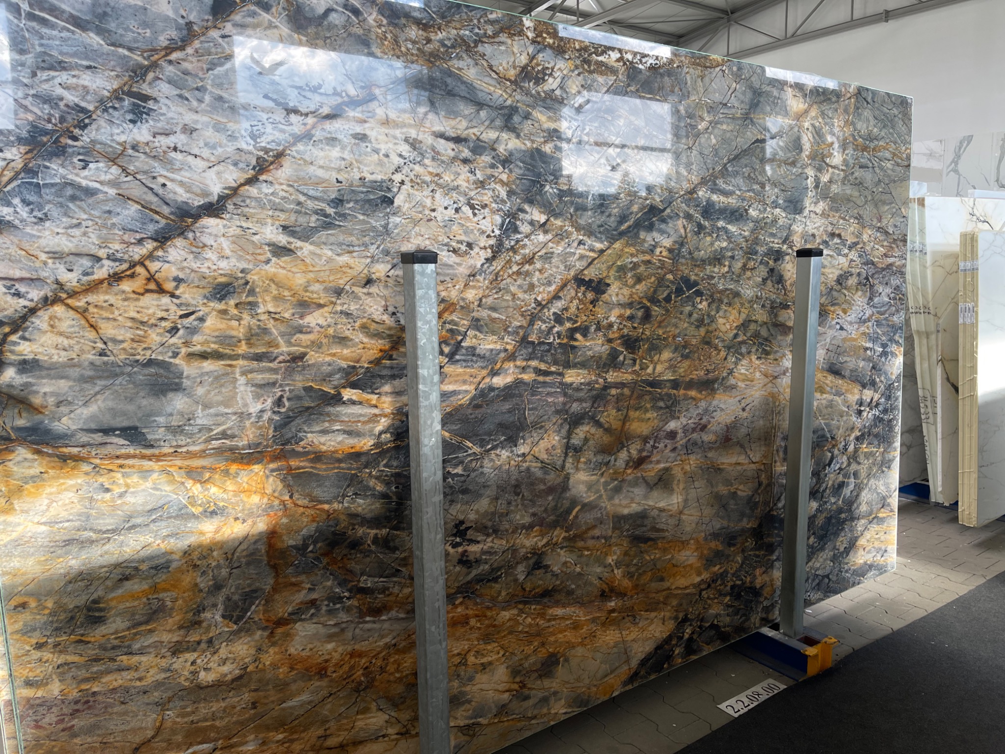 Duża płyta kamienna z wyraźnym, abstrakcyjnym wzorem w odcieniach szarości, bieli i złota, oparta o metalowe podpory w magazynie.