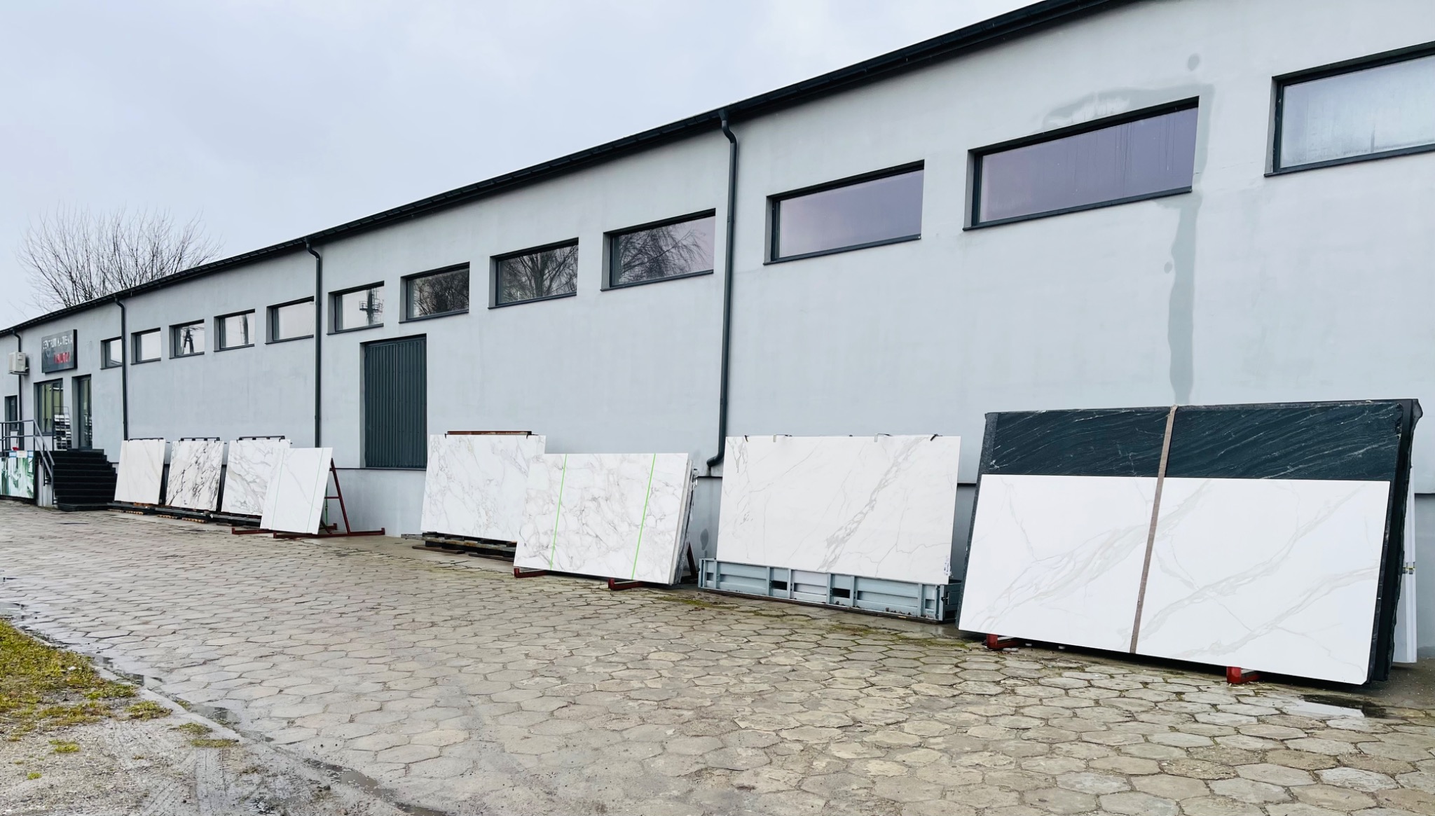 Szeroki kadr na ekspozycję płyt z białego i czarnego konglomeratu na zewnątrz budynku, zabezpieczonych taśmami transportowymi, na tle szarej elewacji z rzędem okien.