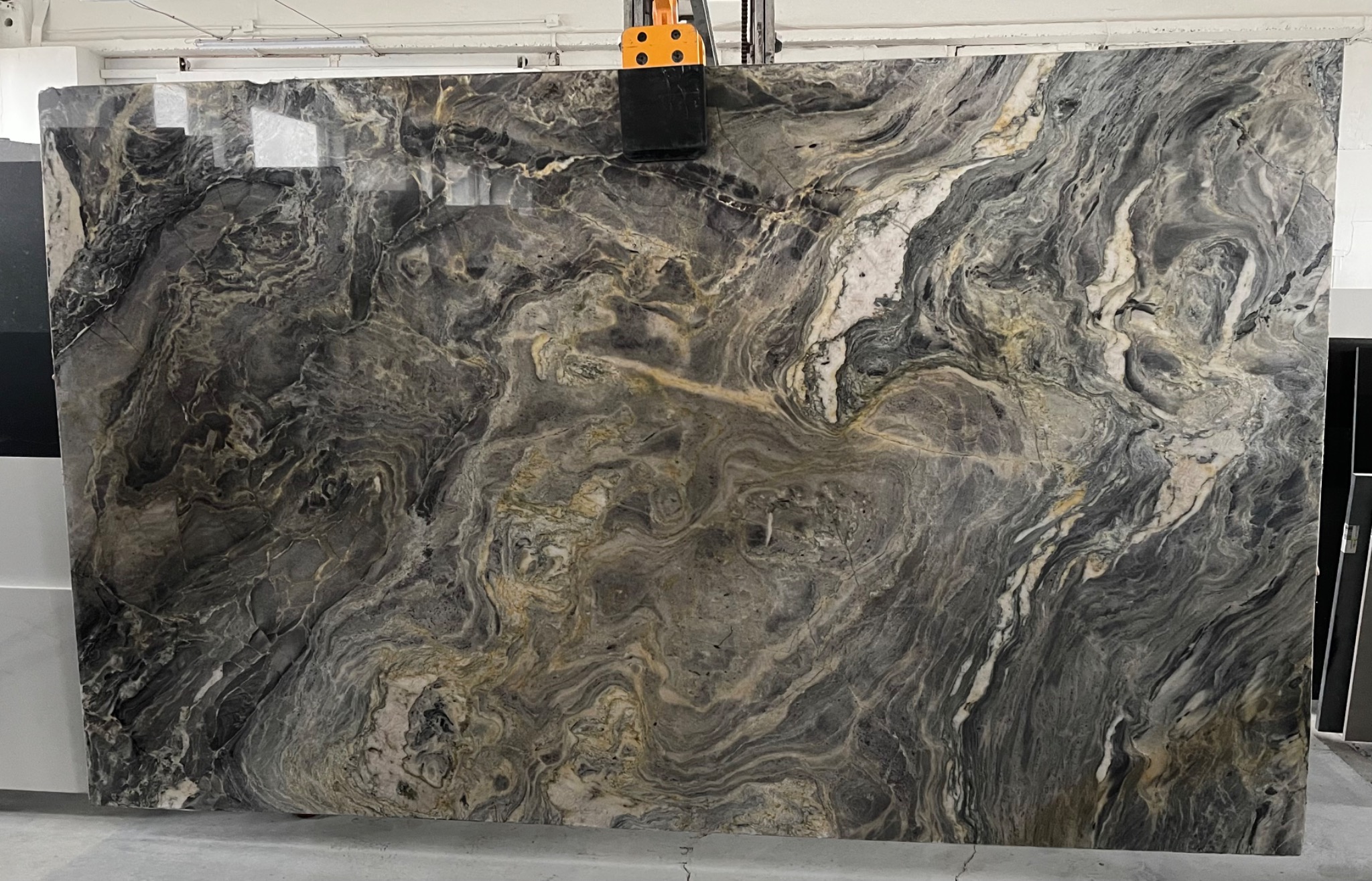Polerowana płyta kwarcytu brazylijskiego o falistej strukturze w odcieniach szarości, bieli i złota, podnoszona przez maszynę w magazynie.
