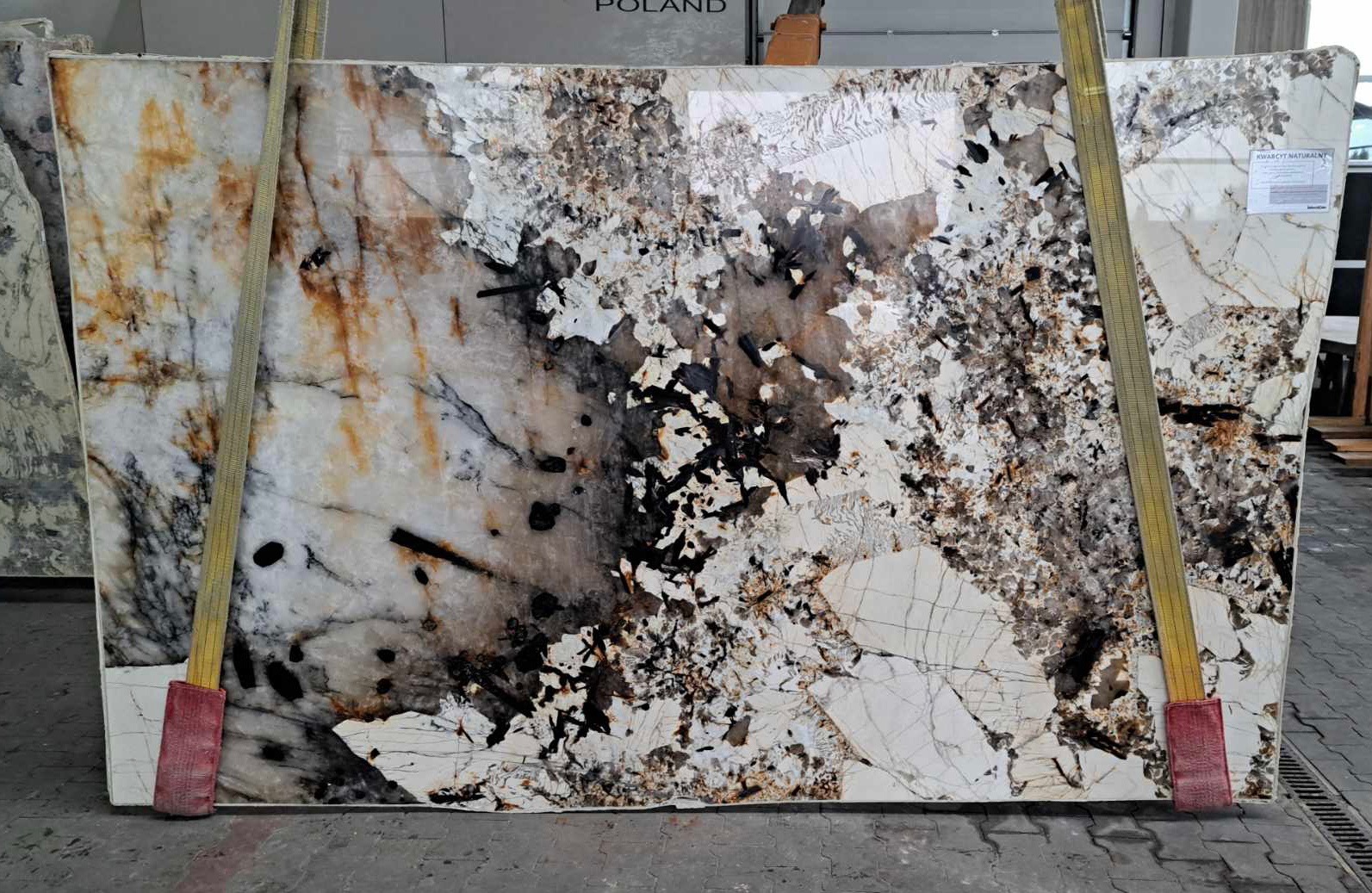 Płyta kwarcytu naturalnego o unikalnym wzorze z przewagą bieli, brązów i czerni, zabezpieczona pasami transportowymi w magazynie.
